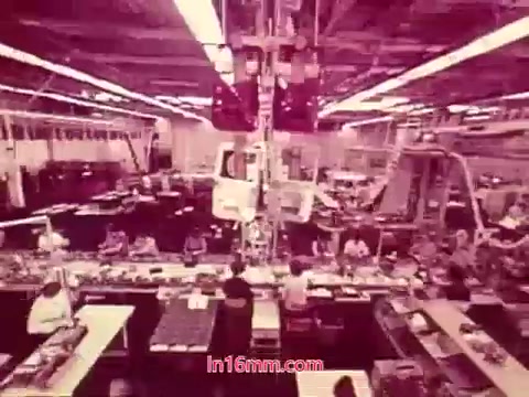 The still from the 16mm film 'Performance' features an industrial setting, likely a factory or assembly line, showcasing a bustling environment. Workers are visible at various stations, engaged in their tasks. The lighting casts a pinkish hue over the scene, adding a unique aesthetic characteristic of that time period. This still represents the film’s focus on industrial processes and teamwork, concluding with credits that detail the production date and the cast and crew involved.