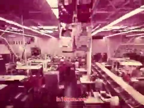 The 16mm still from 'Performance' - Bell System Film (1971) captures a bustling industrial setting, likely a factory or workshop. The scene is filled with machinery and workers engaged in various tasks, conveying a sense of productivity. The lighting is bright and the colors have a muted, vintage tone typical of 16mm prints. This particular frame features the end titles, listing the production date and the cast and crew involved in the film. Overall, it reflects the film's historical context and industrial themes.