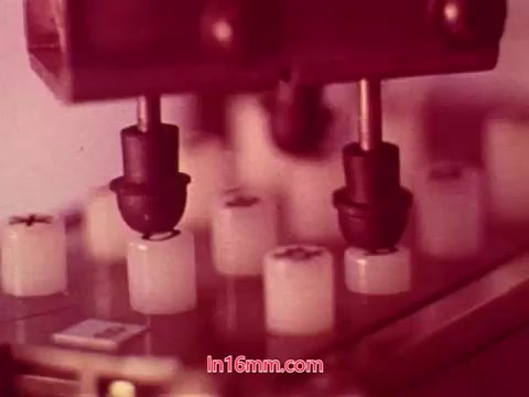 The still from 'Performance' - Bell System Film (1971) showcases a close-up of mechanical components, likely depicting parts of a manufacturing process or machinery. The visual features a series of small, white cylindrical objects positioned on a surface, with apparatus above them, possibly indicating a production line or assembly mechanism. The overall aesthetic is characterized by a vintage color palette, typical of 16mm film, which adds a nostalgic quality to the image. This particular frame likely emphasizes the technical aspects of the film's narrative, connecting to themes of innovation and technology during that era.