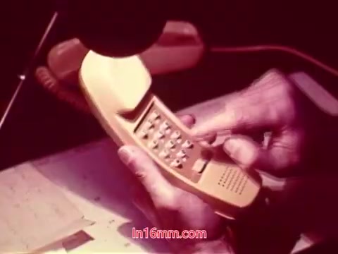 The still from the 16mm film 'Performance' depicts a close-up of hands dialing a yellow telephone, likely a vintage model from the early 1970s. The background suggests a workspace, with papers or documents visible. This scene captures a moment that emphasizes communication, fitting within the film's context as a Bell System production. The end titles, which you mentioned feature the production date and cast and crew, would provide a formal closure to the film, highlighting its historical significance.