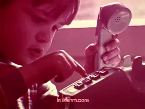 The still from 'Performance' depicts a young child focused on a rotary telephone, pressing buttons with concentration. The warm, soft hues create a nostalgic atmosphere that complements the film's theme. This moment captures the essence of childhood curiosity and communication, showcasing the simplicity of technology from the era. The end titles with production details and credits likely feature after this scene, concluding the film.