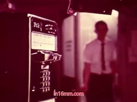 The still from 'Performance,' a Bell System Film from 1971, depicts a man in a white shirt and tie walking towards a payphone. The scene is slightly blurred with a warm color palette, emphasizing the retro aesthetic typical of 16mm prints. This frame likely captures a moment from the film's conclusion, showcasing the end titles that list the production date and credits for the cast and crew. The overall atmosphere hints at a narrative steeped in themes relevant to the era's telecommunications.