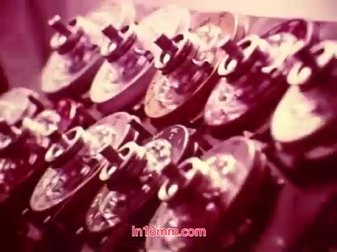 The still from the 16mm film 'Performance' depicts a row of vintage film projectors or film reels, emphasizing the technological aspects of filmmaking. The image likely has a nostalgic tone, captured in a warm color palette. This particular frame likely appears during the end titles segment, providing information about the film's production, including the date, cast, and crew, as it concludes the viewing experience. The film's aesthetic reflects the era of its creation, showcasing a focus on film craft.