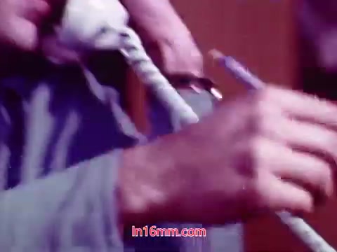 The still from the film 'Performance' shows a close-up of a man's hands holding a telephone receiver, with a pencil or thin object being gripped in the other hand. The focus is on the tactile elements of the scene, emphasizing the connection between technology and communication. The color appears slightly muted, typical of 16mm prints, providing a nostalgic feel. This frame likely contributes to the film's exploration of personal and societal themes from the early '70s.