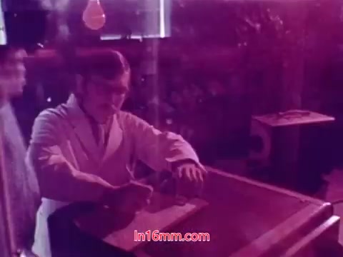 The still from the film 'Performance' features a man in a white lab coat, who appears to be engaged in an activity at a work surface. The image has a pinkish hue, likely due to the film's color processing. In the background, there are shadows and shapes that suggest a laboratory or experimental setting, creating a slightly abstract atmosphere. The overall composition gives a sense of focus on scientific exploration or experimentation, aligning with the film's themes. This still serves as part of the ending credits, providing context about the production and crew involved in the film.