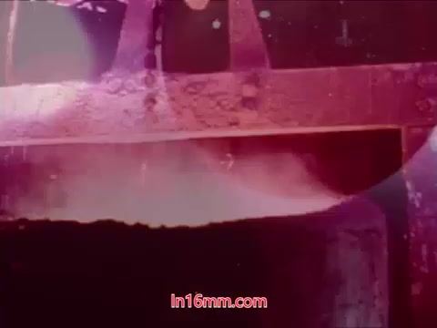 The still from 'Performance' - Bell System Film (1971) features a close-up of industrial machinery, likely a factory setting. The image has a washed-out, colorful aesthetic, with hues of red and pink dominating, suggesting a surreal or abstract portrayal. The focus seems to be on equipment details, with light reflections adding to the visual complexity. The background is slightly blurred, emphasizing the machinery in the foreground. This still represents the film's conclusion, highlighting the cast, crew, and production date in the context of its industrial themes.