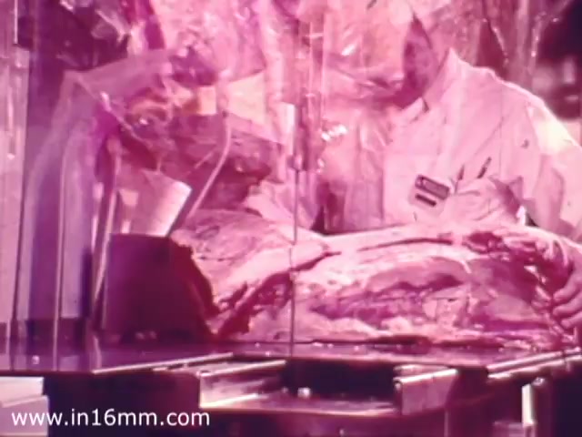 The still features a butcher in a Safeway grocery store, dressed in a white uniform and apron, carefully handling a large cut of meat. The setting is characterized by glass displays and meat packing materials, typical of 1970s grocery presentation. The color tone is warm, giving a nostalgic feel. The focus appears to be on the quality and presentation of the meat, highlighting the store’s commitment to fresh products.