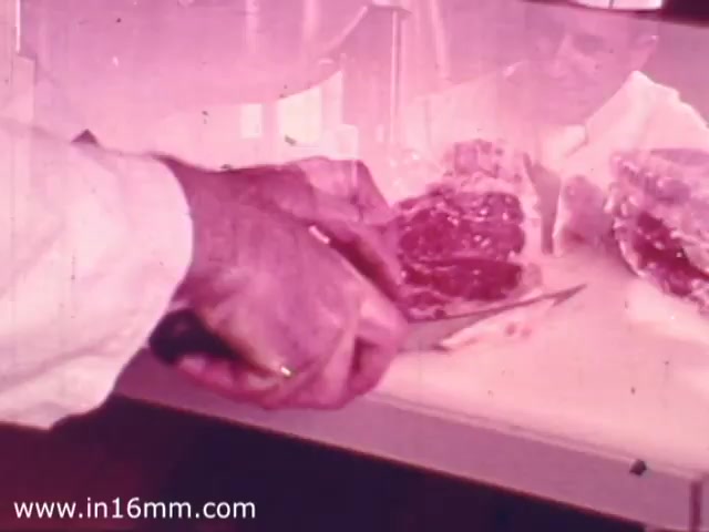 The still features a close-up of a hand wearing a white coat, skillfully cutting through a piece of meat on a cutting board. The scene is likely set in a grocery store context, showcasing the preparation or display of fresh meat products. The color tone has a vintage feel typical of 1970s footage, enhancing the nostalgic aspect of the commercial.