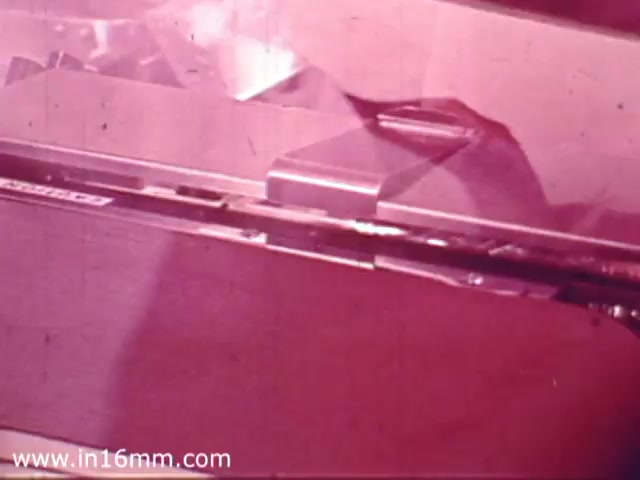 The still from the 1970's Safeway Grocery Store commercial features a close-up view of a checkout counter or cash register. The color is predominantly warm pink, typical of the film's aging process, giving it a vintage feel. Elements like the cash drawer or scanning area are visible, suggesting a focus on the shopping experience. The overall look reflects the aesthetic and service style of grocery stores in that era.