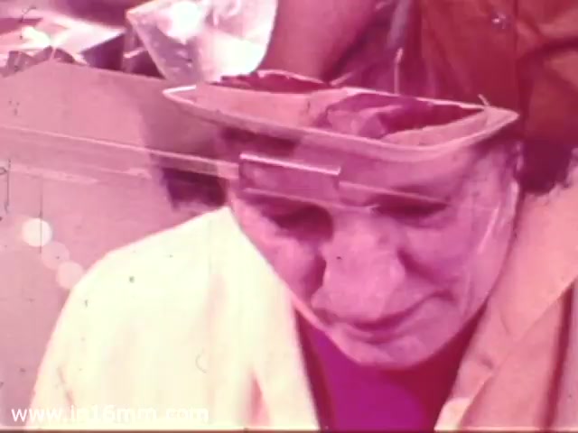 The still depicts a woman in a grocery store setting, likely during a 1970s Safeway commercial. She appears focused or contemplative while wearing a light-colored jacket, and her expression suggests engagement with the products around her. The image has a nostalgic, slightly washed-out aesthetic typical of 16mm film from that era, with visible grain and color distortion.