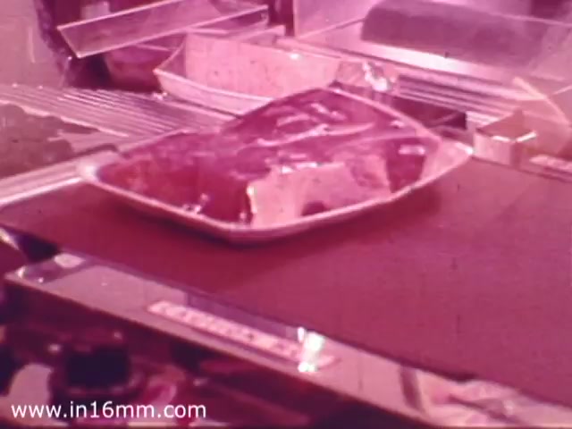 The still depicts a close-up of a meat platter in a 1970s Safeway grocery store commercial. The meat is prominently displayed in a white tray, showcasing its freshness and appealing presentation. The background suggests a typical grocery layout, with various items slightly blurred, emphasizing the focus on the meat product. The overall color tone has a vintage softness, characteristic of 16mm film from that era.
