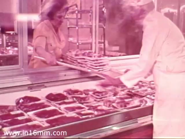The still features a scene inside a 1970s Safeway grocery store, showcasing an employee in a white uniform assisting a woman in a yellow outfit. They are interacting over a display case filled with neatly arranged meat packages. The color tone gives the image a warm, nostalgic feel typical of the era. The commercial likely emphasizes fresh products and customer service.
