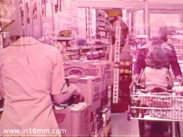 The still captures a scene from a 1970s Safeway grocery store commercial, showcasing a busy supermarket interior. In the foreground, a shopper is seen pushing a cart, while another person is positioned to the side, involved with items on a shelf. The environment is filled with product displays, highlighting the variety of goods available, typical of grocery store settings from that era. The overall color tone has a vintage, slightly faded quality, characteristic of 16mm film prints.