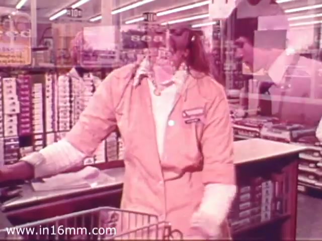 The still captures a scene from a 1970s Safeway grocery store commercial. It features a cashier in a light orange uniform interacting with a customer, while brightly lit aisles filled with grocery products serve as the backdrop. The overall aesthetic reflects the era's marketing style, emphasizing customer service and store variety.