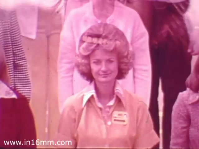 The still shows a young woman wearing a yellow uniform, likely a Safeway employee, with voluminous, styled hair. She is positioned at the forefront of a group, giving a friendly expression. The background consists of other individuals, mostly in casual attire typical of the 1970s. The overall color tone has a warm, slightly faded look, characteristic of 16mm film from that era.