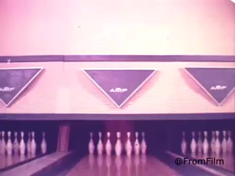 The still depicts a bowling alley, featuring a row of bowling pins set up for a game. The background includes retro-style lane indicators with a triangular design, suggesting a vintage atmosphere. The warm color tones contribute to the nostalgic feel, typical of the 1970s aesthetic. This setting contrasts with the energetic and playful nature of the Kool-Aid Man, emphasizing a fun recreational environment where such commercials may have been featured.