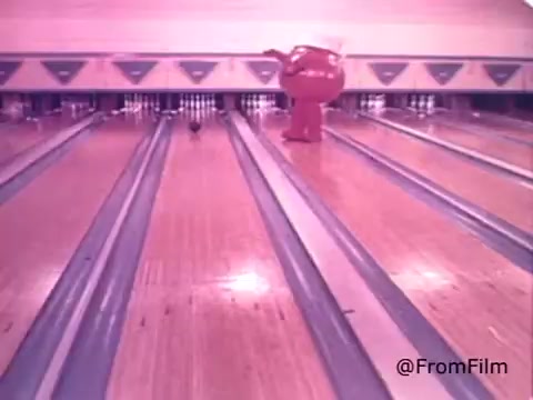 The still captures the Kool-Aid Man, a large, anthropomorphic pitcher filled with cherry Kool-Aid, joyfully navigating a bowling alley. He is prominently positioned in the center, showcasing his playful personality. The wooden floors and bowling lanes form a vibrant backdrop, emphasizing the nostalgic and whimsical tone typical of 1970s commercials. The bright colors and cheerful expression of the Kool-Aid Man convey a sense of fun and excitement, inviting children and viewers to engage with the product.
