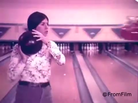 The still depicts a scene from a 1970s Kool-Aid commercial featuring the Kool-Aid Man. In this moment, a young woman is poised to bowl in a bowling alley, holding a bowling ball. The atmosphere is vibrant and playful, capturing the energetic spirit often associated with Kool-Aid and its mascot, the Kool-Aid Man. The setting reflects the leisure activities of the era, emphasizing family and fun.