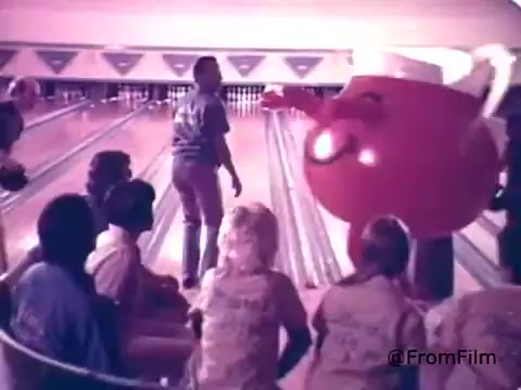 The still depicts the Kool-Aid Man, a large, anthropomorphic pitcher filled with cherry Kool-Aid, engaging with a group of children at a bowling alley. He has a cheerful demeanor and a smiley face, embodying fun and excitement. The setting features bowling lanes and a playful atmosphere, illustrating his role in promoting the Kool-Aid brand as a vibrant and enjoyable drink for kids.