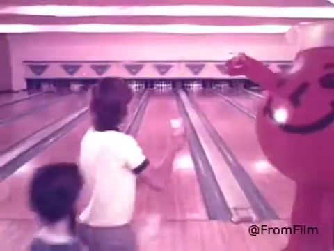 The still depicts the Kool-Aid Man, a large anthropomorphic pitcher filled with cherry Kool-Aid, joyfully engaging with children in a bowling alley setting. The children appear excited, with one child facing the Kool-Aid Man while the other is slightly behind. The bowling lanes stretch out behind them, establishing a fun, vibrant atmosphere typical of 1970s advertising. The cheerful demeanor of the Kool-Aid Man evokes a sense of playfulness and nostalgia.