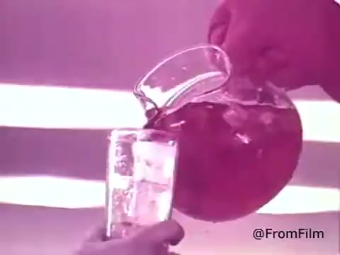 The still features a hand pouring cherry Kool-Aid from a glass pitcher into a clear glass, highlighting the vibrant red color of the drink. The scene evokes a sense of refreshment and fun, typical of 1970s advertisements. The background appears softly lit, enhancing the nostalgic feel of the commercial.