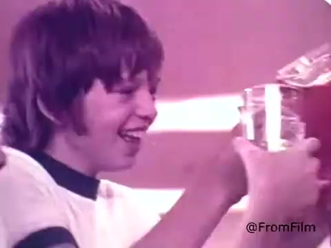 The still features a young boy with a joyful expression, holding up a glass filled with a vibrant, red liquid, likely Kool-Aid. He appears excited, reflecting the fun and energy associated with the Kool-Aid brand. The background has warm, soft lighting, adding to the nostalgic 1970s feel of the scene. The image captures the spirit of children enjoying the drink, highlighting the playful nature of the Kool-Aid Man mascot.