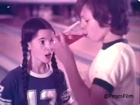 The still from the 1970s Kool-Aid commercial features a scene at a bowling alley. A girl with two braids, wearing a blue shirt with the number 13, looks surprised while a boy in a white and green shirt drinks from a glass filled with bright red Kool-Aid. The setting conveys a fun, youthful energy, highlighting the enjoyment of the drink among children.