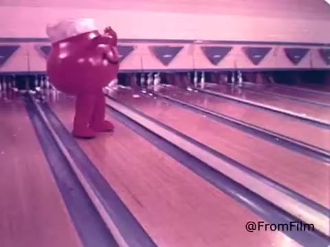 The still features the Kool-Aid Man, a large, anthropomorphic pitcher filled with cherry Kool-Aid, at a bowling alley. He is humorously positioned on the bowling lane, appearing to prepare for a bowling throw. His bright red color and playful demeanor emphasize the character's fun and energetic personality, typical of 1970s advertising aimed at attracting children. The setting hints at an upbeat, recreational environment, aligning with the brand's image of joy and refreshment.
