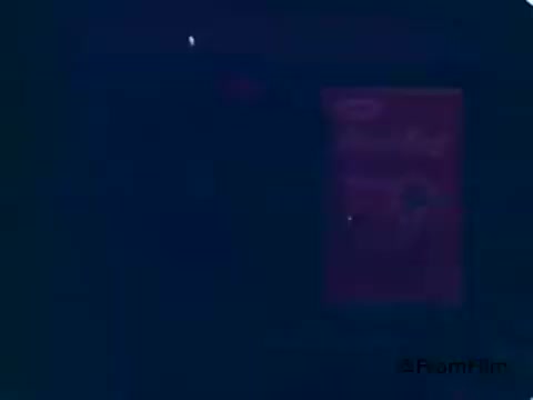 The still from the 1970s Kool-Aid commercial features the Kool-Aid Man, a large, smiling pitcher filled with cherry Kool-Aid. He is joyfully bursting through a wall, embodying a playful and lively spirit. The scene captures the child's excitement and the fun association with the beverage, highlighting the character's role as a friendly, approachable mascot who answers the call of children for a refreshing drink.