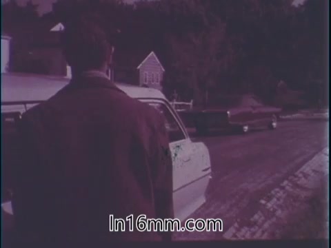 The still from 'Guideposts Magazine PSA - 1960's' depicts a person standing beside a car on a street. The scene conveys a sense of contemplation and responsibility, reflecting the narrator's thoughts about driving safely amidst potential dangers. The muted colors and vintage style emphasize the seriousness of the message, highlighting the importance of being vigilant and prepared while driving. The setting, with houses in the background, adds to the everyday context of the driving experience.