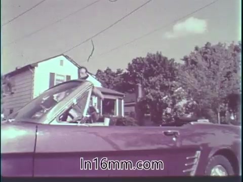 The still from the 'Guideposts Magazine PSA - 1960's' features a man approaching a classic convertible car parked in a suburban setting. The car, likely a 1960s model, is vibrant in color and captures the era's aesthetics. In the background, you can see a modest house and trees, suggesting a residential neighborhood. The context mentions the man's thoughtful approach to driving, emphasizing his awareness of safety and responsibility behind the wheel. This sets a tone of mindfulness and caution, reflective of the PSA's message about the importance of safe driving habits.