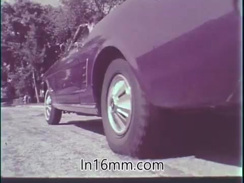 The still shows a vintage car, likely a classic model, positioned in a way that emphasizes its tires and lower body. The surroundings are natural, possibly a park or suburban area, suggesting a sense of calm. The image reflects the themes of responsibility and mindfulness while driving, resonating with the commentary about safety and awareness in traffic. The overall tone is nostalgic, capturing the essence of the 1960s in both aesthetics and messaging.