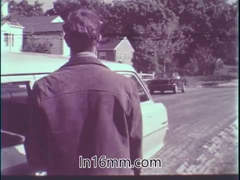 The still from the 'Guideposts Magazine PSA - 1960s' features a man standing beside a parked car, facing the road. He appears thoughtful, possibly contemplating the responsibilities of driving. The background includes trees and houses, indicating a suburban or rural setting. The tone reflects a serious message about safety and mindfulness while driving, aligned with the context of the quote regarding the importance of being a responsible driver.
