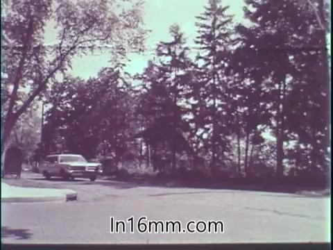The still from the 'Guideposts Magazine PSA - 1960's' features a calm suburban street scene with a white car navigating a corner. Surrounding the road are lush green trees, suggesting a peaceful environment. The imagery ties into a reflective narrative about the responsibility of driving, emphasizing awareness and mindfulness to prevent accidents. The tranquil atmosphere underscores the importance of being vigilant while on the road.