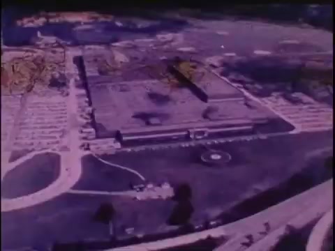 The still from the Sikorsky S-76 promo film depicts an aerial view of a large building, likely a corporate facility or manufacturing site, surrounded by open space and parking lots. The colors appear muted with a pop art aesthetic, giving it a vintage feel typical of the 1970s. The absence of narration adds a layer of mystery, leaving the viewer to interpret the significance of the visuals. This context sets the stage for the eventual introduction of the Sikorsky name, suggesting a focus on innovation or industry within the aviation realm.
