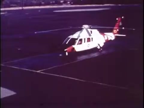 The still from the Sikorsky S-76 promo film features a helicopter on a tarmac, showcasing its sleek design. The aircraft is primarily white with red accents, indicating its modern and professional appearance. The surrounding environment appears to be an airport or helipad. The image captures a moment of readiness, highlighting the helicopter's function and design in a clean, unobtrusive context, typical of promotional material from the late 1970s. Overall, it implies a sense of innovation and reliability without any accompanying narration.