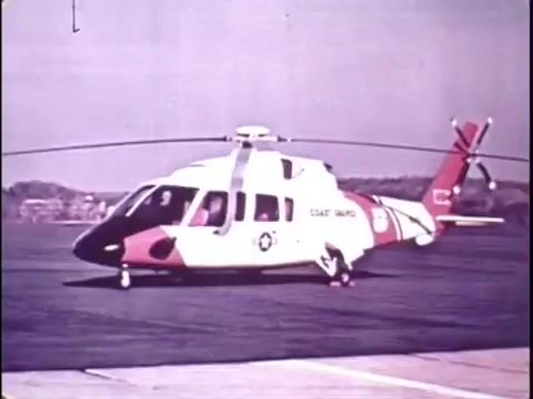 The still captures a Sikorsky S-76 helicopter, prominently marked with 'COAST GUARD' on its side, showcasing its sleek design. The helicopter is positioned on a runway, with its rotor blades in motion, indicating an operational status. The color scheme features a combination of white, red, and black, typical of Coast Guard vehicles. The background hints at an outdoor setting, possibly an airfield or coastal area, emphasizing its readiness for service. The retro aesthetic aligns with the 1977 context of the promo film.