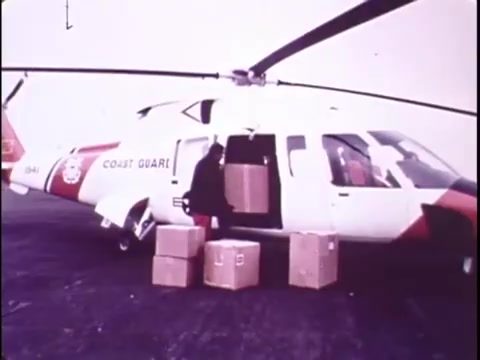 The still from the Sikorsky S-76 promotional film features a Sikorsky S-76 helicopter, prominently marked with the 'COAST GUARD' logo. A person is seen loading cardboard boxes into the helicopter's side door, indicating its utility for transporting cargo. The scene is set against an overcast backdrop, suggesting a practical operational environment. The image captures the helicopter's sleek design and highlights its functional capabilities for missions like those performed by the Coast Guard.