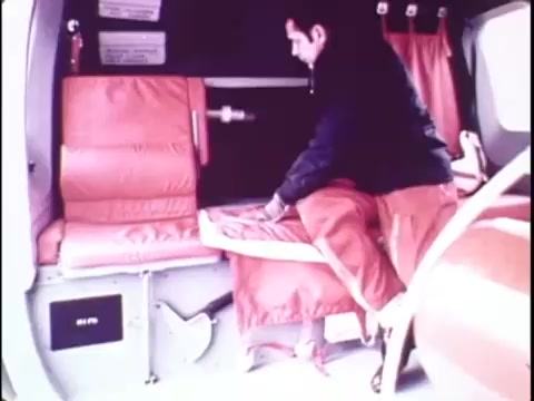 The still from the 1977 Sikorsky S-76 promo film shows a man inside the helicopter, adjusting the red seating. The interior features a practical design, emphasizing functionality. The man appears focused on the seating arrangement, highlighting the attention to comfort and safety in the S-76. The vibrant colors and minimalist setting contribute to a professional and sleek aesthetic typical of the era's promotional material.