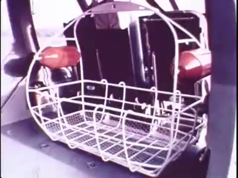 The still from the 1977 Sikorsky S-76 promo film features a close-up of a wire basket with red handles attached. The basket is likely intended for carrying supplies or equipment, emphasizing the utility and functionality of the Sikorsky S-76 helicopter. The image provides a practical glimpse into the design elements, hinting at the helicopter's features aimed at enhancing operational efficiency. The context of a silent workprint means that visual elements must convey the message without narration.