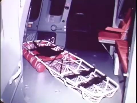 The still from the Sikorsky S-76 promo film features the interior of the helicopter. It showcases a medical stretcher secured in the cabin alongside red seating. The environment is utilitarian, emphasizing practicality and functionality. The color palette is muted, and the lighting suggests a focus on the helicopter's operational features, which are likely highlighted throughout the film. The silent nature of the workprint leaves the visual elements to convey the S-76's design and utility without narration.