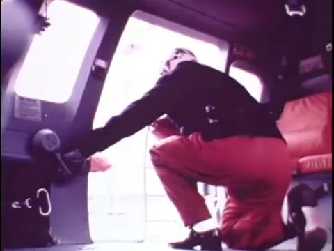 The still from the Sikorsky S-76 promo film features a crew member in red pants and a black jacket, crouching inside the helicopter. The individual's body is partially turned toward the doorway, suggesting they are preparing for takeoff or observing the external environment. The interior is equipped with seating and control elements, showcasing the operational space. The overall aesthetic reflects the era's color palette and design sensibilities.