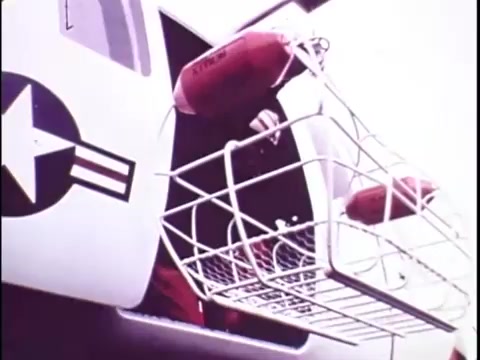 The still from the Sikorsky S-76 promo film shows the side of a helicopter featuring a distinctive design with a star emblem and military stripes. An individual is depicted near the open door, interacting with a basket-like contraption that holds what appears to be several red cylindrical objects, possibly flotation devices. The overall setting suggests a focus on operational versatility, likely related to rescue or transport missions. The silent nature of the film leaves the imagery open to interpretation without accompanying narration.