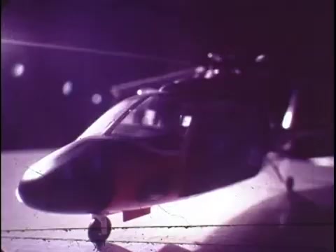 The still from the 1977 Sikorsky S-76 promo film captures a model of the helicopter, showcasing its sleek design under dramatic lighting. The focus is on the helicopter's profile, highlighting its aerodynamic features and modern aesthetic typical of the era. The background likely has a blurred effect, emphasizing the model as the central subject. The overall tone gives a sense of innovation and style associated with the Sikorsky brand, though the absence of narration leaves the viewer curious about its specific features and advantages.