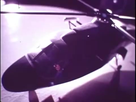The still features a Sikorsky S-76 helicopter, showcasing its sleek design and features. The helicopter is viewed from above, emphasizing the rotor blades and cockpit. The lighting highlights the contours of the aircraft, creating a polished, professional appearance that aligns with typical promotional materials. This image likely serves to capture the viewer's attention, drawing focus to the advanced engineering of the S-76 in the context of its promotional film from the late 1970s.