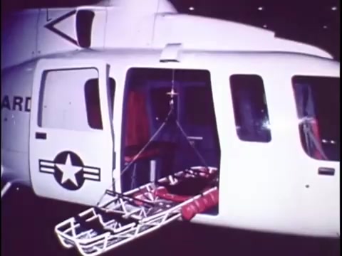 The still features a Sikorsky S-76 helicopter, showcasing its open cargo door. Inside, there's a stretcher hanging, indicating its potential use for medical or rescue operations. The helicopter bears military markings, including a star emblem. The overall aesthetic reflects the promotional character of the film, emphasizing the helicopter's functionality and design. The image captures a moment that suggests readiness for critical missions, despite the lack of accompanying narration.