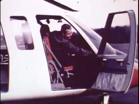 The still from the 1977 Sikorsky S-76 promo film shows a pilot seated in the cockpit of the helicopter, appearing to reach for the door or control. The interior features red seats and a modern design for its time, indicating comfort and functionality. The lighting suggests an outdoor setting, possibly during a demonstration or test flight. This captures a moment of anticipation and readiness, highlighting the aircraft’s design and pilot ergonomics.