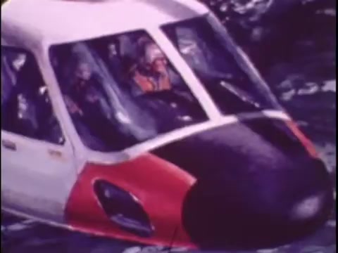 The still from the 1977 Sikorsky S-76 promo film features the helicopter close-up, showcasing its distinctive design. The cockpit area is prominent, highlighting the sleek lines and the contrasting color scheme of white and red with a black nose. The image likely captures pilots inside, emphasizing the operational aspect of the aircraft. The overall tone is professional, aimed at demonstrating the capabilities and appeal of the Sikorsky S-76.