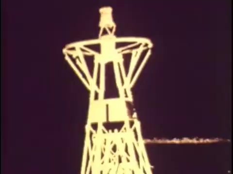 The still from the 'Sikorsky S-76 promo film from 1977' depicts a stark, industrial structure resembling a radio or microwave tower, prominently featuring sharp lines and geometric shapes. The image has a vintage aesthetic, with a muted color palette typical of the era's film. The absence of sound makes the visual elements stand out, focusing attention on the structure, possibly symbolizing technology and communication. This imagery aligns with the broader promotional theme of innovation and advancement in aviation, setting the stage for the Sikorsky S-76 helicopter introduction that follows.