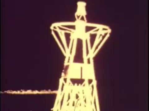 The still from the 1977 Sikorsky S-76 promotional film depicts a vintage oil rig structure, likely illuminated at night. The bright outlines emphasize its towering presence, which may suggest industrial or technological themes. Given the context of the film, this image could signify the advancements in aviation and energy industries, connecting the Sikorsky S-76 to modern technologies of the era. The absence of sound makes it challenging to grasp specific messaging, but the visual focus could evoke a sense of innovation and progress.